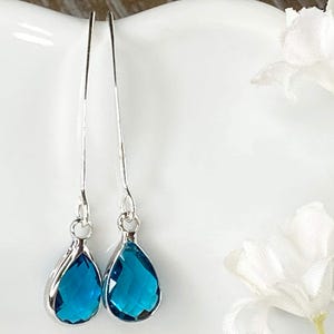 May include: A pair of silver-toned drop earrings featuring faceted, pear-shaped blue glass stones. The earrings have long, slender silver-toned wires that extend upward from the teardrop-shaped settings, creating a sleek and elongated profile.