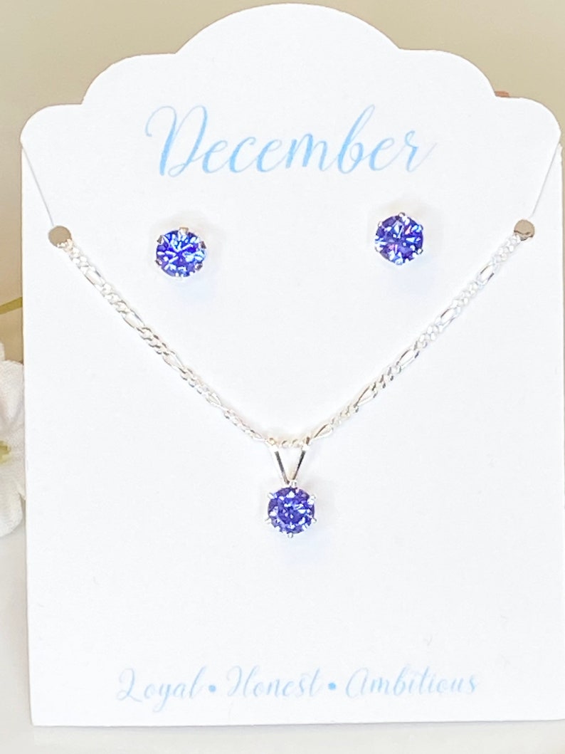 Puede incluir: Un conjunto de collar y pendientes de plata con una piedra preciosa azul. La tarjeta dice "Diciembre, Leal, Honesto, Ambicioso".