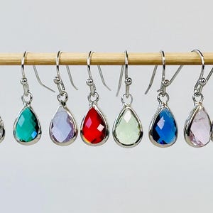 May include: A collection of teardrop-shaped earrings in various colors, including red, purple, blue, and yellow, are displayed on a wooden dowel. Each earring has a silver-colored hook.