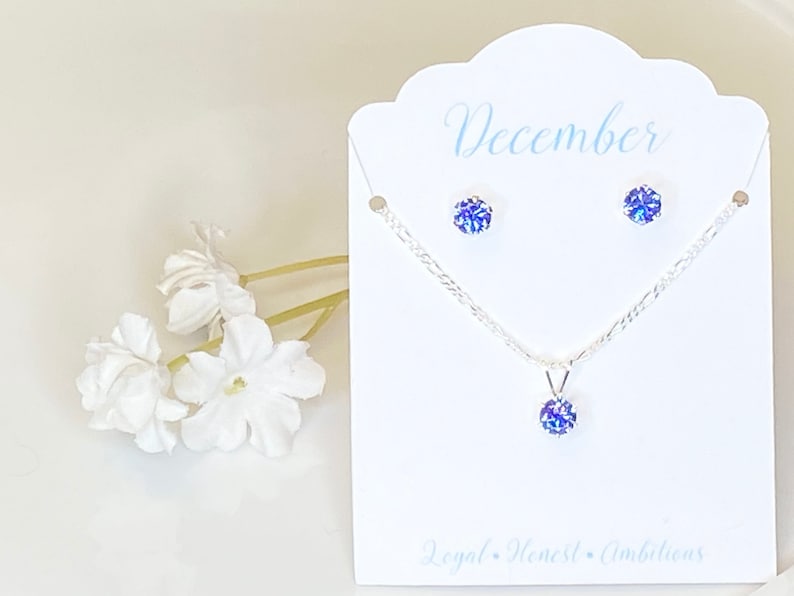 Puede incluir: Un conjunto de collar y pendientes de plata con un colgante de piedra preciosa azul y pendientes a juego. La tarjeta dice "Diciembre" y "Leal, honesto, ambicioso".