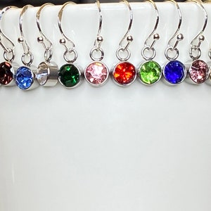 May include: A collection of silver-toned dangle earrings, each featuring a different colored round gemstone. The gemstones include red, purple, blue, clear, green, pink, orange, lime green, dark blue, light pink, yellow, and turquoise. The earrings are displayed against a white background.