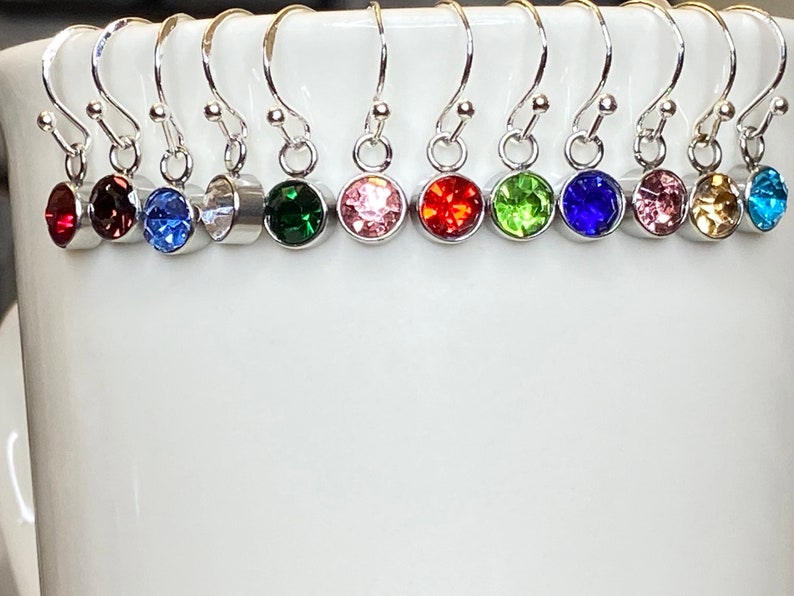 May include: A collection of silver-toned dangle earrings, each featuring a different colored round gemstone. The gemstones include red, purple, blue, clear, green, pink, orange, lime green, dark blue, light pink, yellow, and turquoise. The earrings are displayed against a white background.