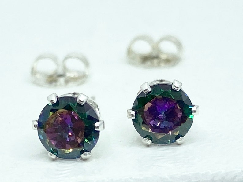 Puede incluir: Un par de pendientes de plata con piedras preciosas redondas e iridiscentes. Las piedras preciosas tienen una base verde oscuro con destellos de morado, azul y rojo.