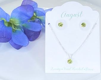 May include: A silver-toned necklace and matching stud earrings set featuring round, light green peridot-style stones. The jewelry is displayed on a white card printed with the text August, Loving, Kind-Hearted, Brave in light green script.