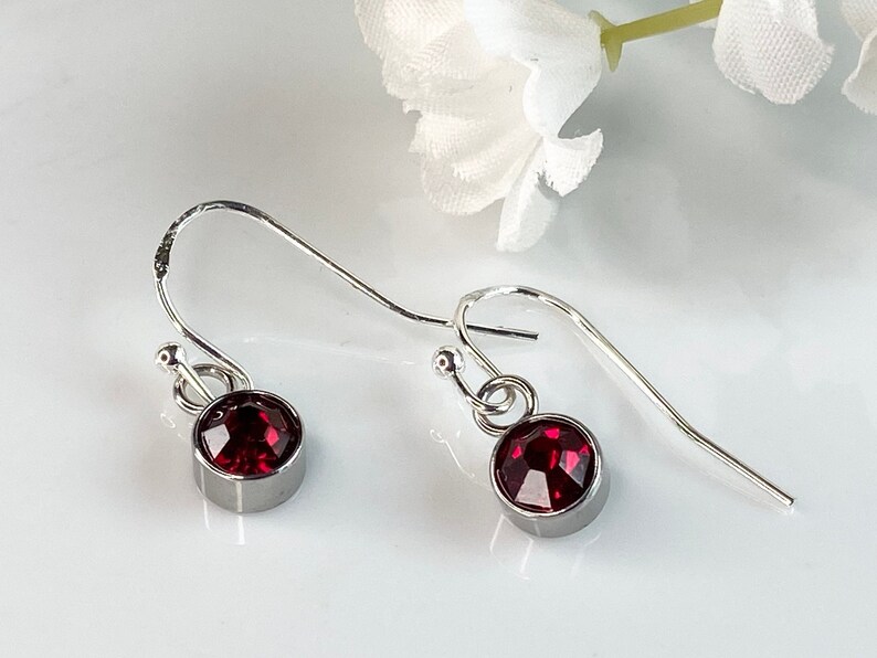 May include: A pair of silver-tone earrings with a deep red, round gemstone set in a bezel. The earrings have a simple hook design, perfect for everyday wear. The gemstones are approximately 0.25 inches in diameter.