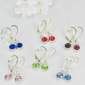 May include: A collection of lever-back earrings with colorful, round gemstones. The earrings are silver-toned and feature gemstones in various colors, including red, blue, green, pink, and purple. The earrings are displayed on a white surface with white flowers in the background.