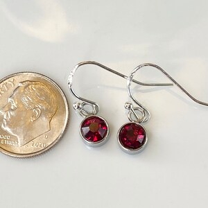 May include: A pair of silver-toned drop earrings with round, deep red gemstones. The earrings are shown next to a U.S. dime for size comparison. The gemstones are set in a bezel setting, and the earrings have a simple, elegant design.