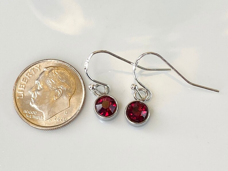May include: A pair of silver-toned drop earrings with round, deep red gemstones. The earrings are shown next to a U.S. dime for size comparison. The gemstones are set in a bezel setting, and the earrings have a simple, elegant design.