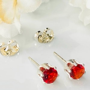 May include: A pair of stud earrings featuring round, faceted, vibrant red stones set in silver-toned metal prongs. Each earring includes a matching silver-toned metal butterfly-style back closure, positioned separately from the main earring posts.