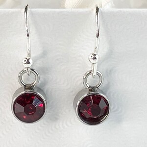 May include: A pair of dangle earrings featuring a deep red, faceted gemstone set in a silver-colored bezel. The earrings have a simple design with a hook closure and a small, round accent above the gemstone. The gemstones are approximately 0.5 inches in diameter.