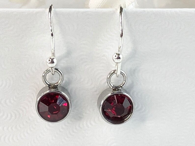 May include: A pair of dangle earrings featuring a deep red, faceted gemstone set in a silver-colored bezel. The earrings have a simple design with a hook closure and a small, round accent above the gemstone. The gemstones are approximately 0.5 inches in diameter.