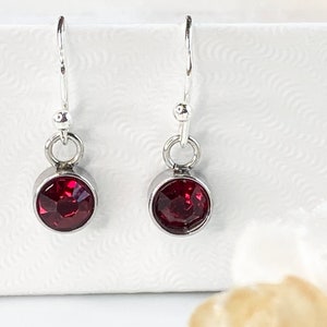 May include: A pair of dangle earrings with a silver-tone metal setting. Each earring features a round, faceted, deep red gemstone. The earrings have a simple design with a hook closure. The gemstones are approximately 0.3 inches in diameter.