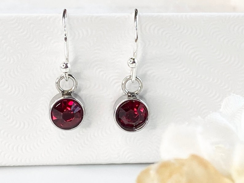 May include: A pair of dangle earrings with a silver-tone metal setting. Each earring features a round, faceted, deep red gemstone. The earrings have a simple design with a hook closure. The gemstones are approximately 0.3 inches in diameter.