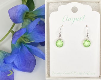 May include: Green peridot gemstone dangle earrings in sterling silver. The earrings are displayed in a white box with the word "August" and the phrase "Loving - Kind-Hearted - Brave".