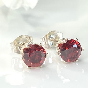 May include: A pair of stud earrings featuring round, faceted deep red stones set in a silver-toned metal prong setting. Each earring includes a matching silver-toned metal butterfly back closure, showcasing a classic design with a polished finish.