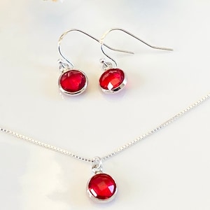 May include: A jewelry set consisting of a necklace and a pair of earrings. Each piece features a round, faceted, deep red stone set in a polished silver-toned bezel. The necklace includes a delicate silver-toned box chain, and the earrings have hooks.