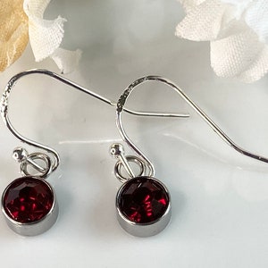 May include: A pair of silver-toned dangle earrings. Each earring features a round, faceted, deep red gemstone set in a bezel. The earrings have a simple, elegant design with a hook closure.