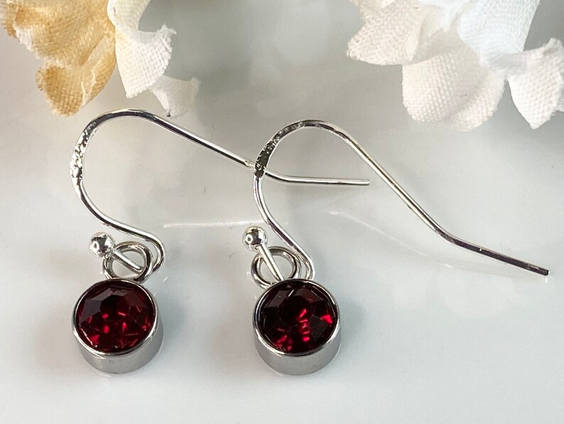 May include: A pair of silver-toned dangle earrings. Each earring features a round, faceted, deep red gemstone set in a bezel. The earrings have a simple, elegant design with a hook closure.