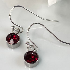 May include: A pair of silver-toned drop earrings. Each earring features a round, faceted, deep red gemstone set in a bezel. The earrings have a simple, elegant design with a hook closure.