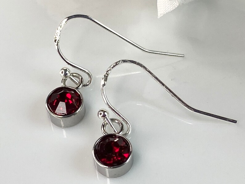 May include: A pair of silver-toned drop earrings. Each earring features a round, faceted, deep red gemstone set in a bezel. The earrings have a simple, elegant design with a hook closure.