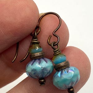 Könnte beinhalten: Handgefertigte Ohrringe mit türkisfarbenen und blauen Glasperlen. Die Ohrringe haben kupferfarbene Haken und dekorative Elemente. Die Perlen haben ein facettiertes Design und sind mit kleineren Perlen und Metallbeschlägen akzentuiert.