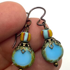 Puede incluir: Un par de pendientes hechos a mano con cuentas azules claras, amarillas y rojas. Los pendientes tienen una cuenta de vidrio azul claro en forma de lágrima con una tapa de metal decorativa y una cuenta rayada más pequeña. Los ganchos para las orejas son de metal oscuro.