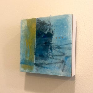Può includere: Un'opera d'arte da parete quadrata con una composizione astratta a strati. La superficie presenta una fascia verticale di vernice dorata testurizzata sulla sinistra, in contrasto con velature blu scuro e chiaro, linee di carboncino e tre piccoli punti dorati.