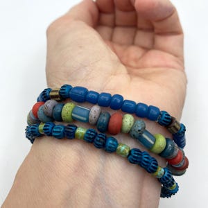 Peut inclure: Trois bracelets de perles aux tons de bleu, rouge, vert et beige sont empilés sur un poignet. Les bracelets présentent une variété de formes et de tailles de perles, créant un accessoire texturé et coloré. Les bracelets sont un bijou unique et accrocheur.