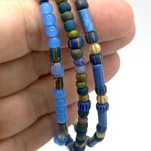 以下が含まれることがあります： 青、緑、クリーム色のビーズブレスレット3本。ブレスレットは、丸型、円筒型、模様入りのビーズなど、さまざまな形状とサイズのビーズが特徴です。白い背景に、手に持ったブレスレット。