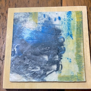 Può includere: Un'opera d'arte quadrata caratterizzata da una composizione strutturata e stratificata di vernice grigio scuro, blu e giallo pallido su una superficie piana, montata su una base quadrata in legno chiaro più grande con venature visibili.