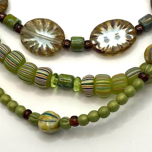 Puede incluir: Primer plano de tres collares de cuentas. El collar superior presenta cuentas grandes, ovaladas e iridiscentes. El collar del medio tiene cuentas verdes y rayadas. El collar inferior tiene cuentas pequeñas, redondas y verdes y una sola cuenta estampada.