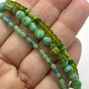 以下が含まれることがあります： 緑と青の色合いのビーズブレスレット3本。ブレスレットは、丸いビーズ、ストライプビーズ、ファセットビーズなど、さまざまなサイズのビーズと形状が特徴です。カラーパレットには、明るい緑と濃い緑、ターコイズ、金色のタッチが含まれています。