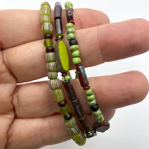 Op de afbeelding: Drie armbanden met een verscheidenheid aan kleurrijke kralen. De armbanden hebben groene, rode, bruine en gele kralen. De armbanden worden in een hand gehouden.