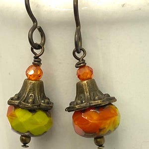 May include: A pair of handcrafted earrings with a vintage aesthetic. Each earring features a bronze-toned metal cap and a faceted, colorful bead. One bead is yellow and green, the other is orange and yellow. The earrings have bronze-toned ear wires.
