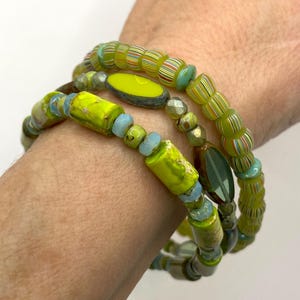May include: Three beaded bracelets in shades of green, yellow, and blue. The bracelets feature a variety of bead shapes, including cylindrical, oval, and faceted beads. The beads have striped, speckled, and solid patterns.