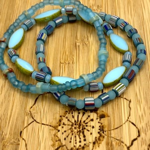 以下が含まれることがあります： 青と白の色合いのビーズネックレス3連。ネックレスは、小さな丸いビーズと、より大きな楕円形のビーズが混ざっています。一部のビーズにはカラフルなストライプがあります。ネックレスは木製の表面に配置されています。
