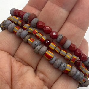 May include: A multi-strand bracelet featuring a combination of faceted deep red beads, textured grey disc-shaped beads, and grey beads with yellow and red striped patterns. The beads are strung together in a repeating sequence to form the bracelet.