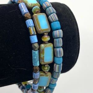 Puede incluir: Tres pulseras de cuentas en tonos azules, turquesas y marrones. Las pulseras presentan una variedad de formas de cuentas, incluyendo rectangulares, cilíndricas y redondas. Algunas cuentas tienen un patrón de rayas. Las pulseras están apiladas juntas.