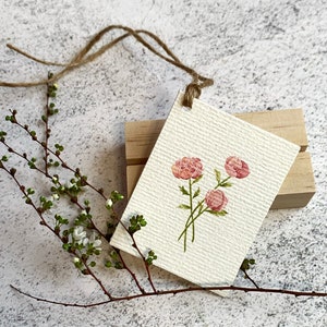 以下が含まれることがあります： 水彩画のタグ。3つのピンク色の牡丹の花と緑色の茎が描かれています。タグはテクスチャ加工された紙でできており、ひもで木のブロックに取り付けられています。小さな緑の葉と白い花が付いた枝が前景にあります。