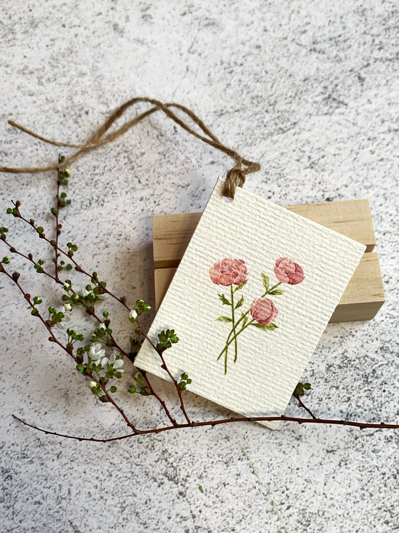 以下が含まれることがあります： 水彩画のタグ。3つのピンク色の牡丹の花と緑色の茎が描かれています。タグはテクスチャ加工された紙でできており、ひもで木のブロックに取り付けられています。小さな緑の葉と白い花が付いた枝が前景にあります。