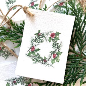 May include: White gift tags with a watercolor holly wreath design. The wreath features green leaves and red berries. The tags are tied with twine and surrounded by green cedar branches, creating a festive holiday theme.