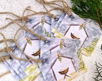 May include: A stack of square Christmas gift tags with watercolor illustrations of a manger scene. Each tag is connected with twine. The artwork features a manger, a star, and a tree, set against a gray and white sky.
