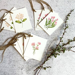 以下が含まれることがあります： ピンクの花と緑の葉の水彩イラストが描かれた、花のギフトタグのコレクション。各タグは長方形で、表面はざらざらしており、吊り下げ用のジュートの紐が付いています。緑の小枝が横に置かれています。