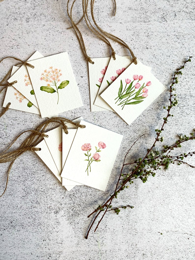 以下が含まれることがあります： ピンクの花と緑の葉の水彩イラストが描かれた、花のギフトタグのコレクション。各タグは長方形で、表面はざらざらしており、吊り下げ用のジュートの紐が付いています。緑の小枝が横に置かれています。