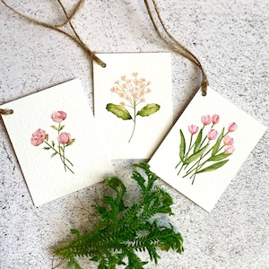 以下が含まれることがあります： 紐のハンガーが付いた、3つの水彩画の花のアートタグ。各タグには、ピンクのバラ、繊細な白と桃色の花、ピンクのチューリップなど、異なる花が描かれています。緑の葉の小枝がタグの下にあります。