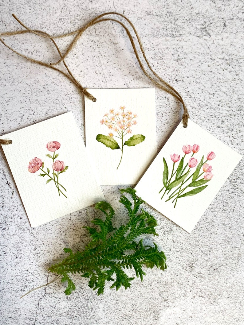 以下が含まれることがあります： 紐のハンガーが付いた、3つの水彩画の花のアートタグ。各タグには、ピンクのバラ、繊細な白と桃色の花、ピンクのチューリップなど、異なる花が描かれています。緑の葉の小枝がタグの下にあります。