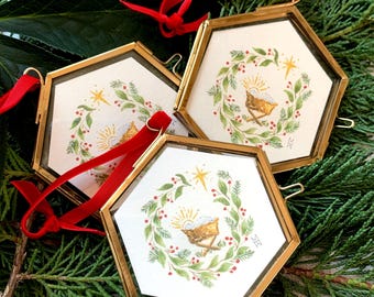 May include: Three hexagonal Christmas ornaments with gold frames and red velvet ribbons. Each ornament features a watercolor illustration of a nativity scene within a green wreath with red berries. The ornaments rest on a bed of green foliage.