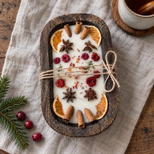 Puede incluir: Una vela en un cuenco de madera rectangular relleno de cera blanca, decorado con rodajas de naranja seca, ramas de canela, anís estrellado y bayas rojas. El cuenco está envuelto con cuerda natural atada en un lazo a la derecha.