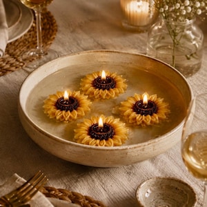 Puede incluir: Un cuenco de cerámica lleno de agua, con cuatro velas encendidas con forma de girasol. Las velas tienen pétalos amarillos y centros de color marrón oscuro. La escena está ambientada en una mesa con copas de vino y arreglos florales.