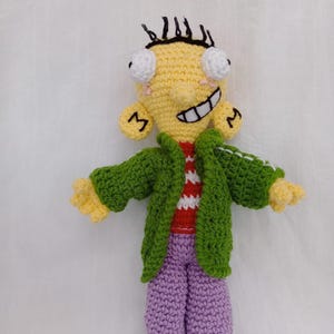 Puede incluir: Muñeca de ganchillo hecha a mano de un personaje de dibujos animados. La muñeca tiene una cabeza amarilla con pelo negro, ojos blancos grandes y una amplia sonrisa. Viste una chaqueta verde, una camisa a rayas rojas y blancas, pantalones morados y zapatos blancos y negros.